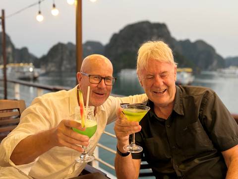       Two men raise bright green cocktails on a cruise deck with Halong Bay karst islands behind
  