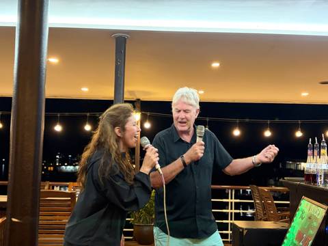       Man and woman enthusiastically singing karaoke on an outdoor deck lit by string lights
  