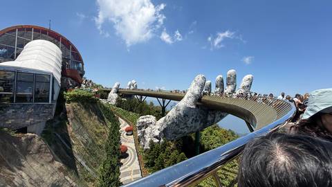       Crowds walk the dramatic Golden Bridge held by giant stone hands above forested mountains
  