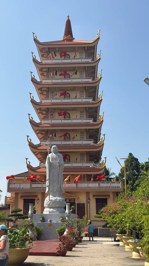       Tall tiered pagoda with a standing Buddha statue in front against clear blue sky
  