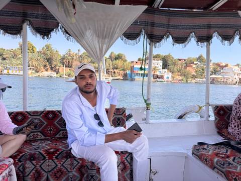       Man in white attire sits aboard a shaded felucca with colorful village on Nile bank behind
  