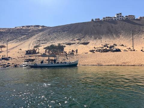       Small boat approaches sandy desert riverbank where camels and handlers wait
  