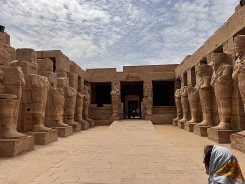       Row of colossal statues lining courtyard entrance at Karnak Temple under partly cloudy sky
  