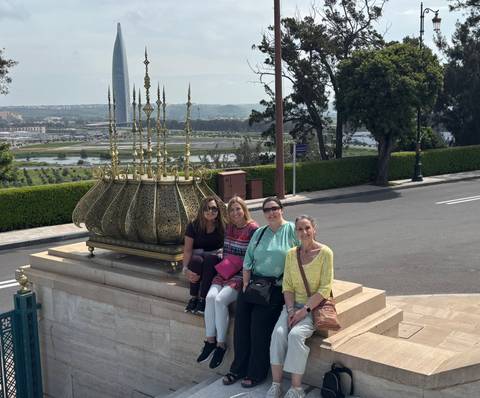       Four women sit smiling in front of an ornate golden incense holder with a cityscape in the background.
  