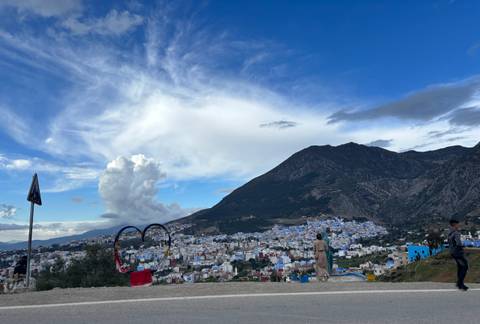       Panoramic lookout over the blue-washed town of Chefchaouen nestled below a dramatic mountain under a big sky.
  