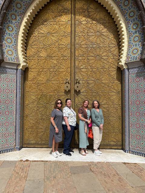       Four women stand smiling in front of ornate golden doors of the Royal Palace in Fes.
  