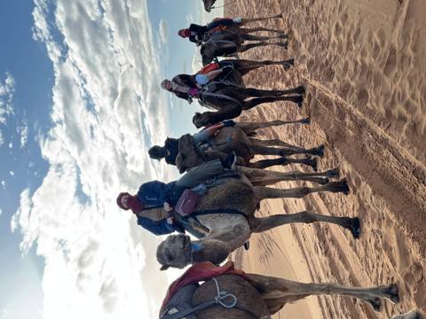       Line of travelers riding camels across reddish Sahara sand dunes under a partly cloudy sky.
  