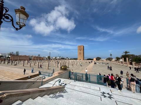       Wide plaza with marble columns and Hassan Tower in Rabat with tourists scattered around under blue skies.
  