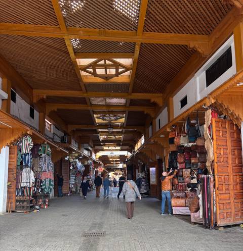       Covered souk lined with leather bags and textiles, wooden ceiling beams and shoppers walking through.
  