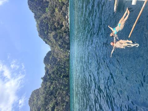       Two women float on crystal-clear blue water surrounded by steep forested limestone cliffs.
  