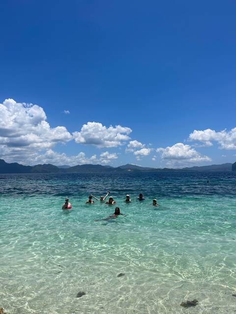       Group of travelers waist-deep in turquoise sea waving toward the camera with mountains on the horizon.
  