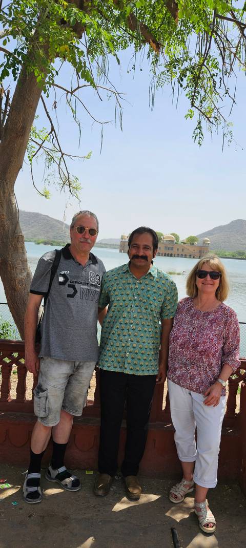       Three people pose lakeside with Jaipur’s Jal Mahal palace and arid hills behind them.
  