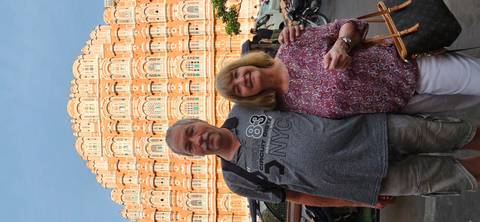       Couple smiles before the intricate pink facade of Hawa Mahal in Jaipur on a sunny day.
  
