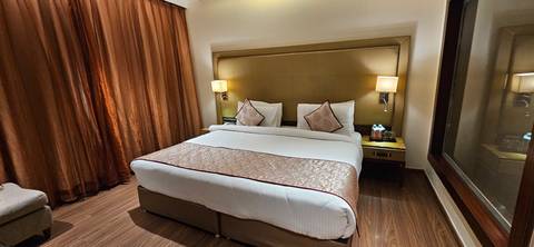       Modern hotel room with king bed, warm lighting, brown wood floors and draped curtains.
  