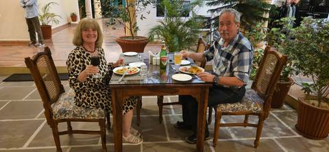       Couple enjoys dinner at a wooden table in a plant-filled restaurant, smiling toward the camera.
  