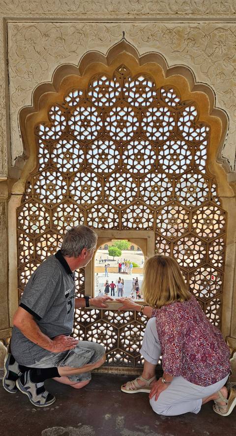       Older tourists look through an ornate lattice window at a palace courtyard with bright sunshine outside.
  