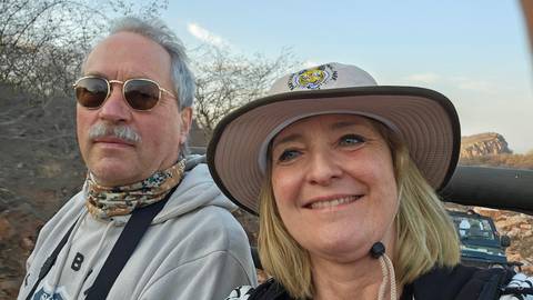       Selfie of an older couple in safari gear with rocky scrubland and leafless bushes behind them.
  