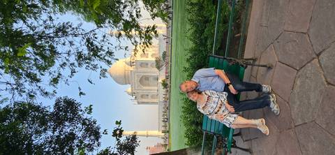       Couple sitting on a bench with the Taj Mahal framed by morning light and green lawns.
  