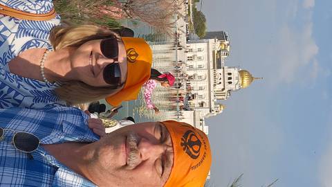       Couple wearing orange headscarves smiles in front of the white marble Sikh temple and reflecting pool.
  