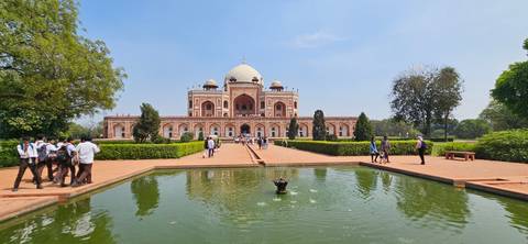       Wide view of Humayun’s Tomb reflected in a tranquil pool, framed by manicured gardens and blue skies.
  