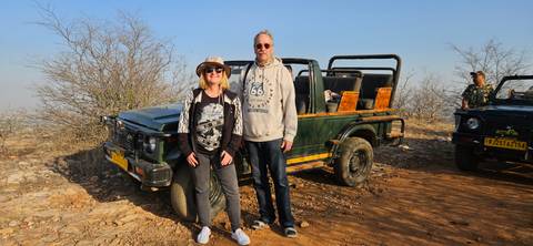       Couple in front of an open-top safari jeep on a dusty track surrounded by dry scrub.
  