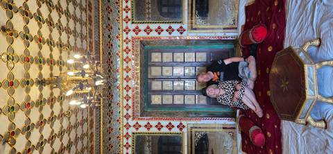       Couple seated on rich red cushions in an opulent palace room decorated with mirrors and chandeliers.
  