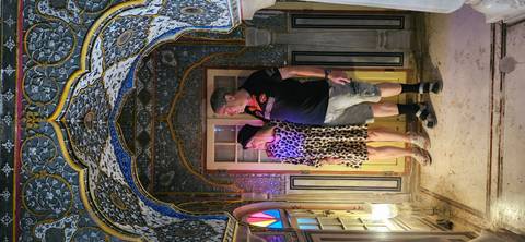       Couple admire colorful stained-glass windows beneath an ornate painted archway inside a palace chamber.
  