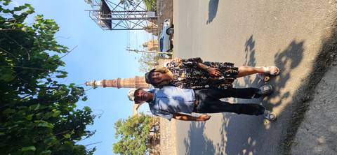      Couple poses on roadside with Qutub Minar rising behind against a clear blue Delhi sky.
  