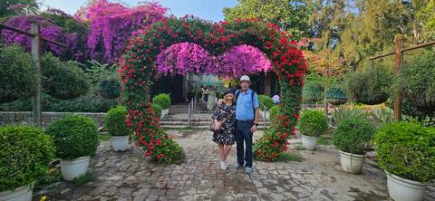       Couple under a heart-shaped arch of red and pink flowers in a lush garden walkway.
  