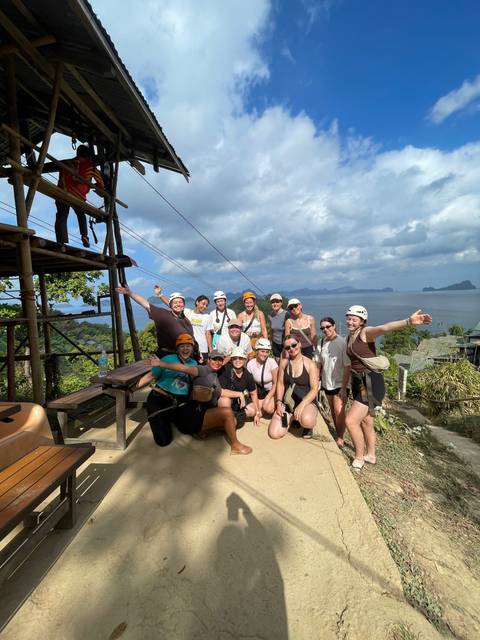       Group wearing helmets celebrates atop a coastal viewpoint with islands and sea behind.
  