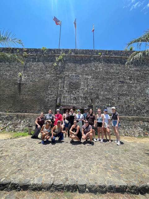       Tour group poses in front of a massive stone fort wall under harsh midday sun.
  