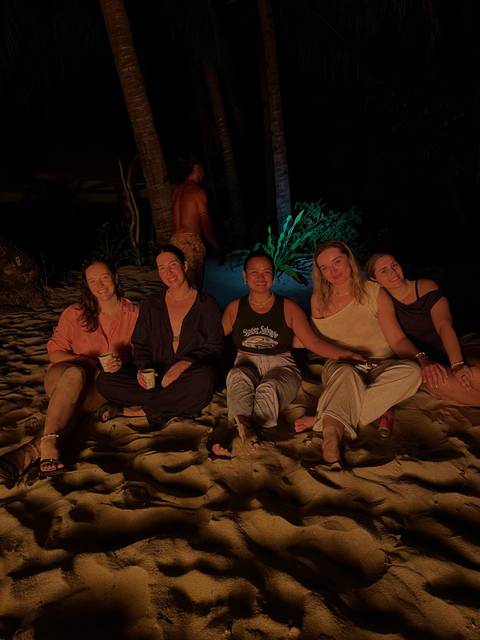       Group of friends sit on a beach around a fire, warmly lit against the dark night background.
  