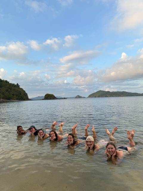       Laughing travelers in the sea raise their feet above the water with island scenery beyond.
  