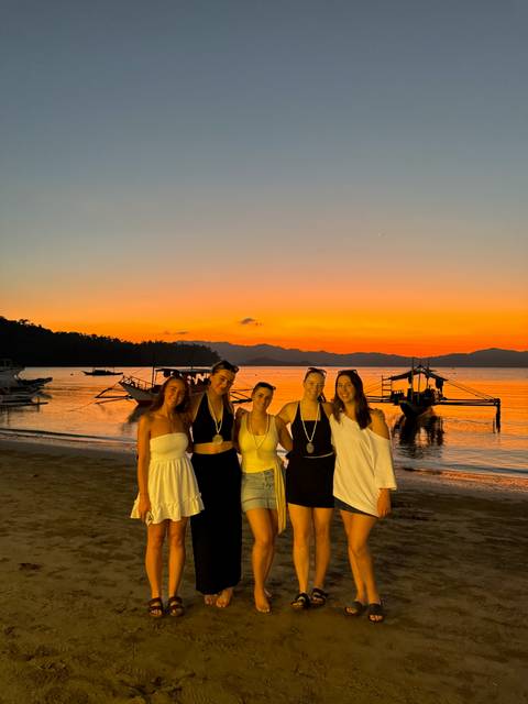       Five women pose on the beach at vivid orange sunset with boats and mountains in the distance.
  