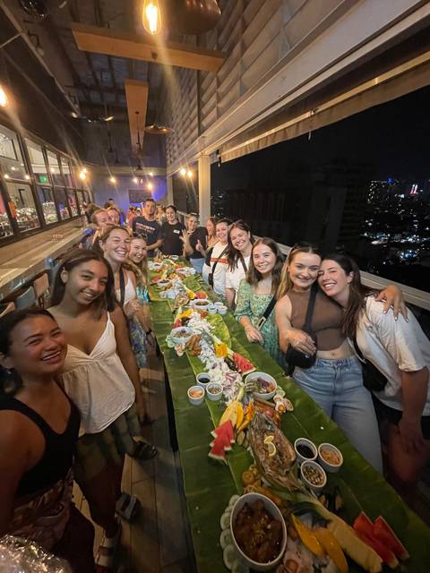      Long table feast on a rooftop at night with a large group enjoying Filipino boodle-fight dinner.
  