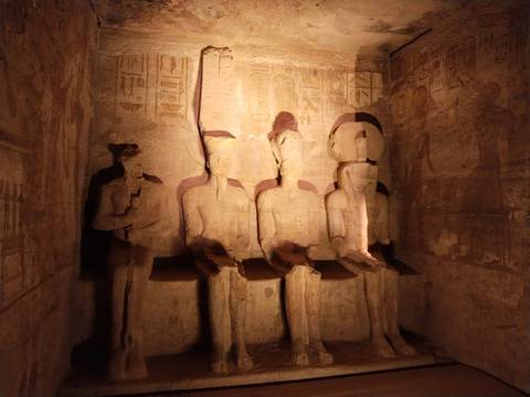       Dimly lit interior of Abu Simbel shows four seated pharaonic statues carved from stone.
  