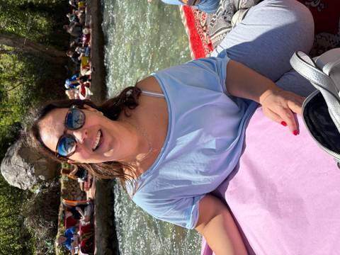       Smiling woman in sunglasses relaxing on a riverside cushion with flowing water behind her.
  