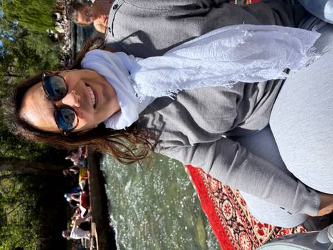       Woman in scarf and sunglasses sitting on a rug by a fast-flowing mountain stream.
  