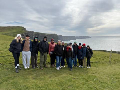       Travel group poses on grassy cliff edge with the Cliffs of Moher behind.
  