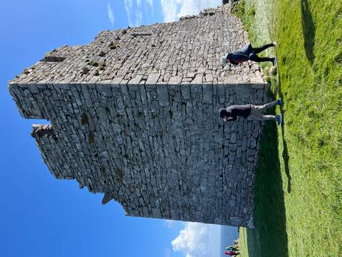      Two travelers walk beside a tall medieval stone tower under clear sky.
  