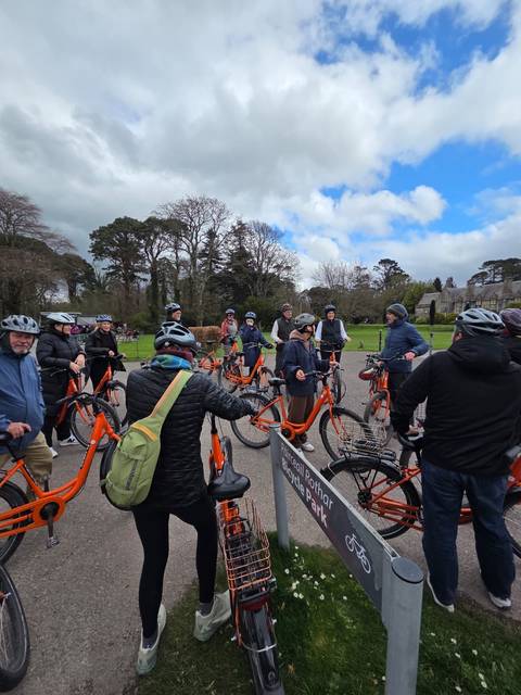       Cycling group gathers with orange bikes in a park beside a manor.
  