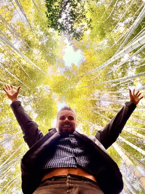       A smiling traveler stretches arms upward beneath towering bamboo stalks.
  
