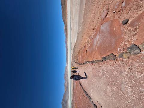       Hikers walk along a red-earth trail toward a wide white salt flat under a deep blue sky.
  