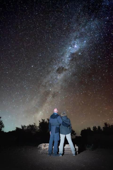       Two stargazers admire the Milky Way glittering across a clear desert night sky.
  