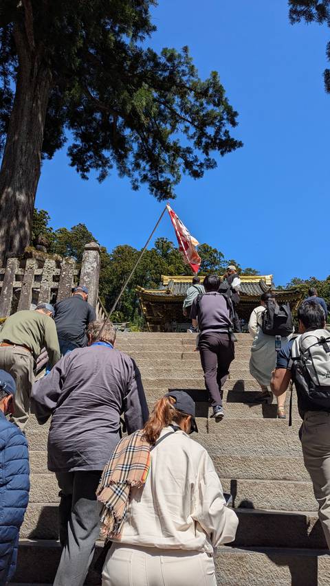       Visitors ascending stone steps toward a shrine roof while a guide holds a flag under a clear blue sky.
  
