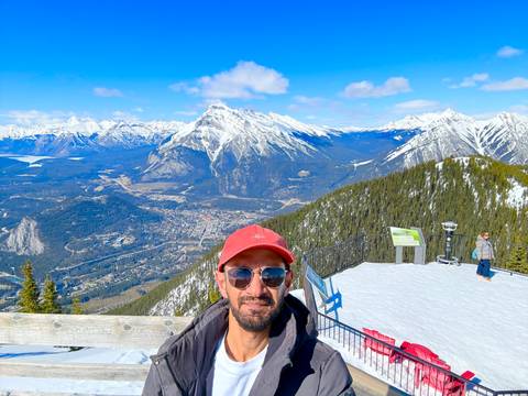       A man in a red cap poses on a snowy observation deck overlooking Banff townsite and dramatic snow-capped peaks.
  