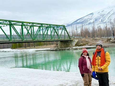       Two travelers stand beside a turquoise river under a green steel bridge with snowy mountains rising behind.
  