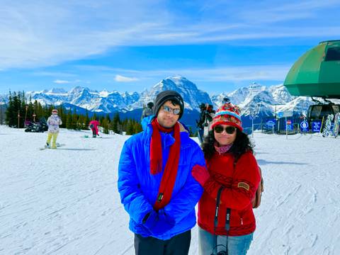       A young pair dressed in bright ski gear pose on a snowy ski field with impressive Rocky Mountain peaks in the background.
  