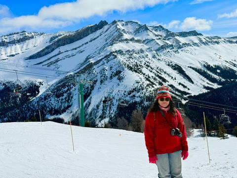       A woman in red stands on a ski slope with chairlifts and striking snowy mountain ridges behind her.
  