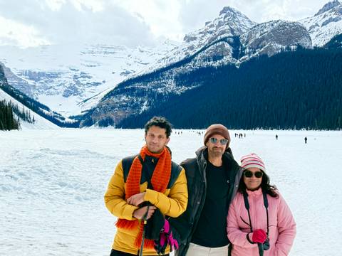       Three family members stand on a frozen lake plain with dramatic snow-covered slopes and evergreen forests behind.
  